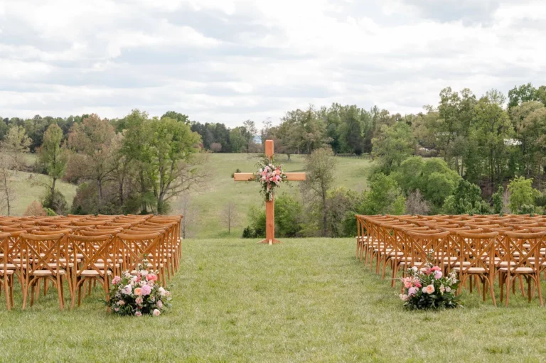 Rows of wooden chairs are arranged outdoors on a grassy field at a Tiffin barn wedding, facing a large wooden cross decorated with pink flowers. Bouquets of pink and white flowers line the aisle, with trees and open fields in the background.