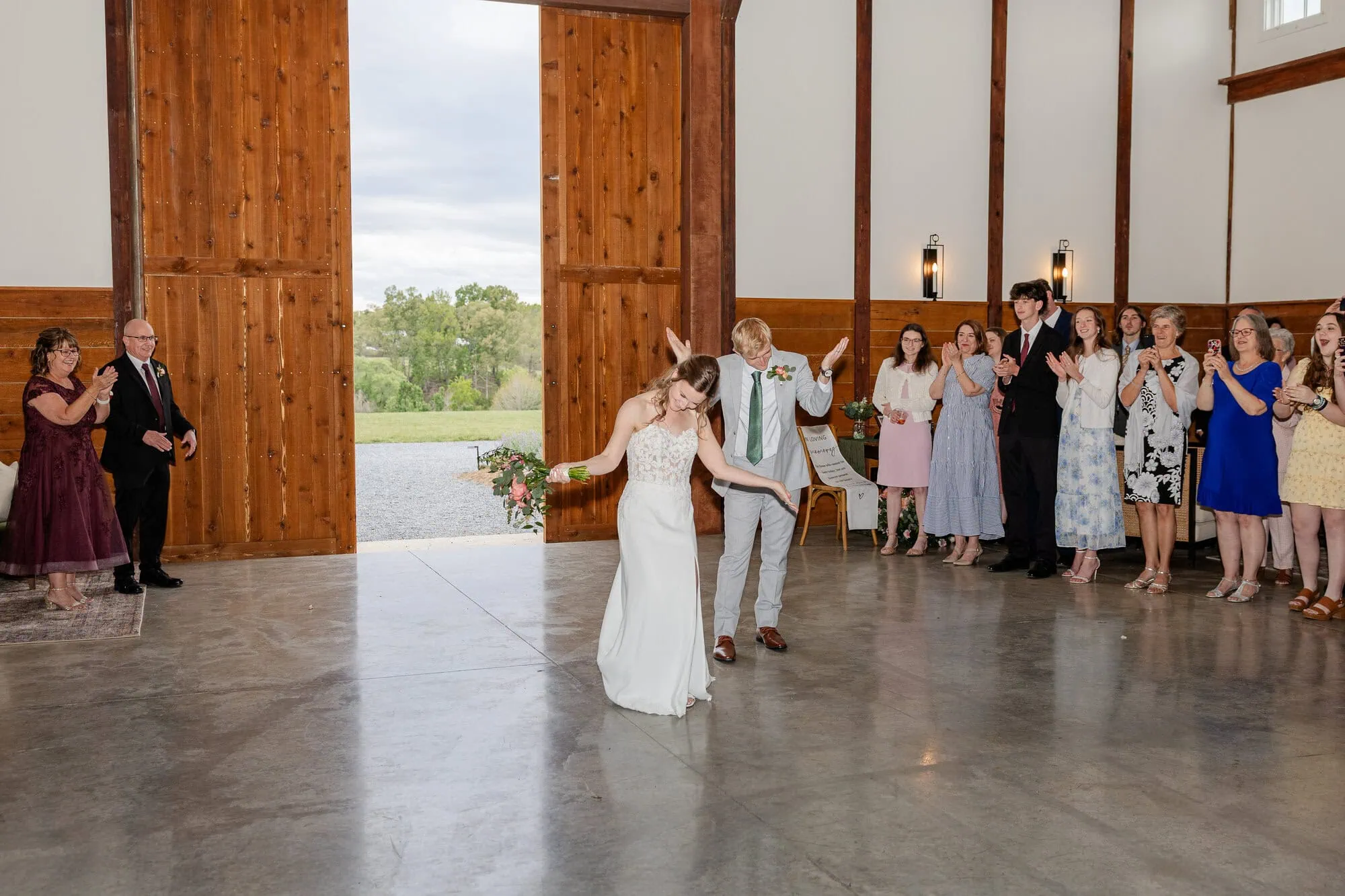 A bride and groom dance joyfully in a rustic Tiffin Barn venue with large open wooden doors, while wedding guests smile, watch, and clap from the side—capturing the magic of a Tiffin Barn wedding.