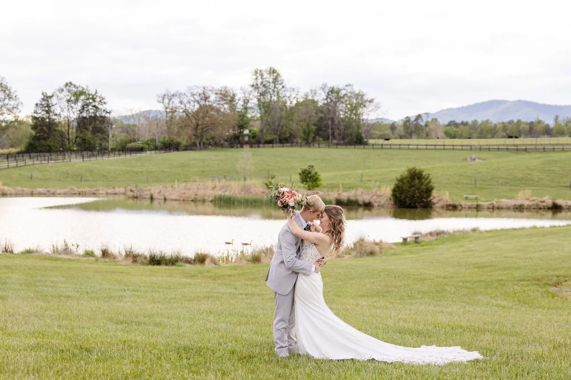 A bride and groom embrace and kiss on a grassy lawn near a pond at their Tiffin barn wedding, with trees and distant mountains in the background. The bride holds a bouquet and lifts one leg as the groom supports her.