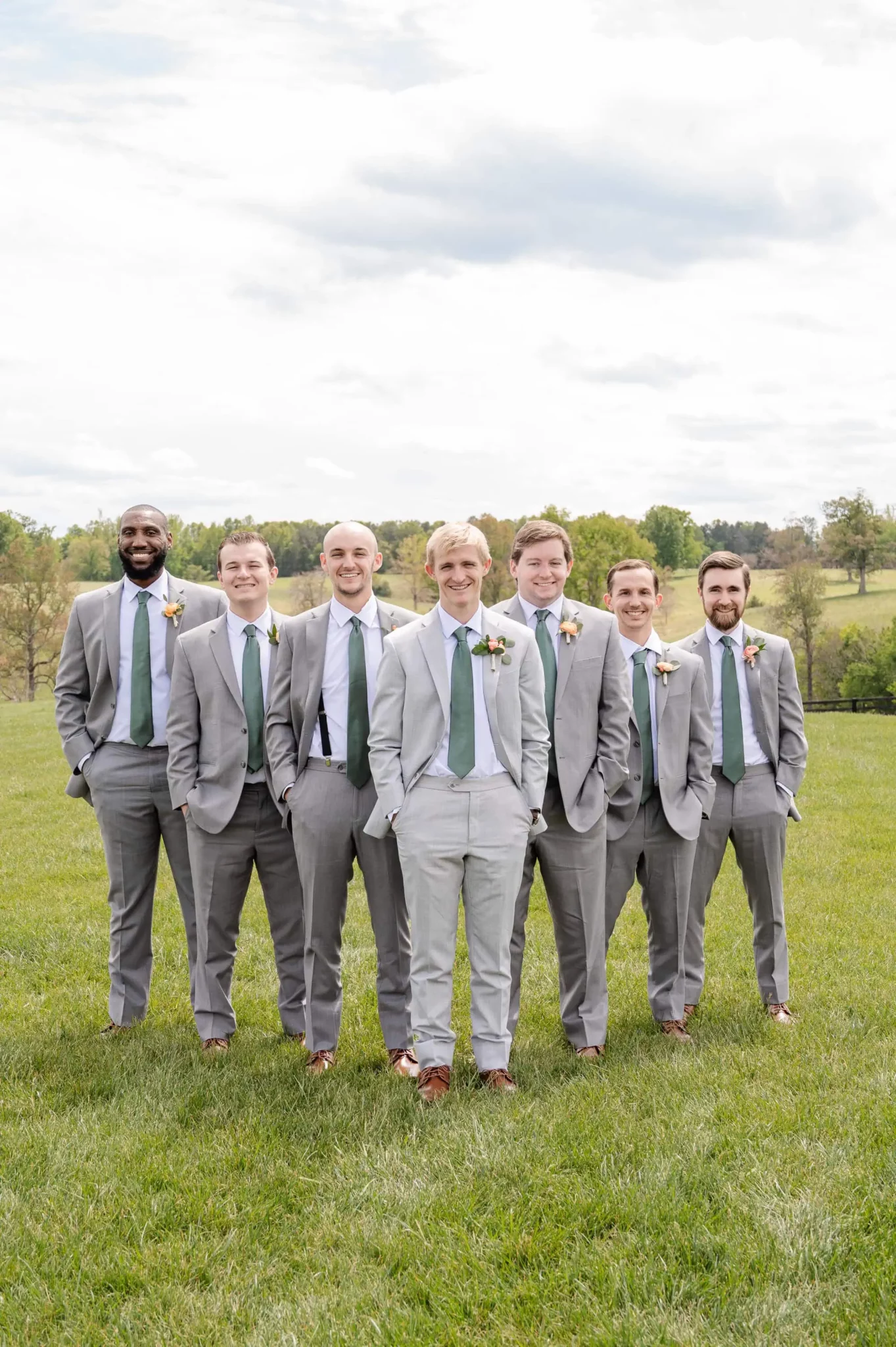 Seven men in light gray suits with green ties and boutonnieres stand on grass, smiling for tiffin barn photos. Behind them, a scenic, tree-filled landscape stretches under a cloudy sky—perfect for a Tiffin barn wedding.