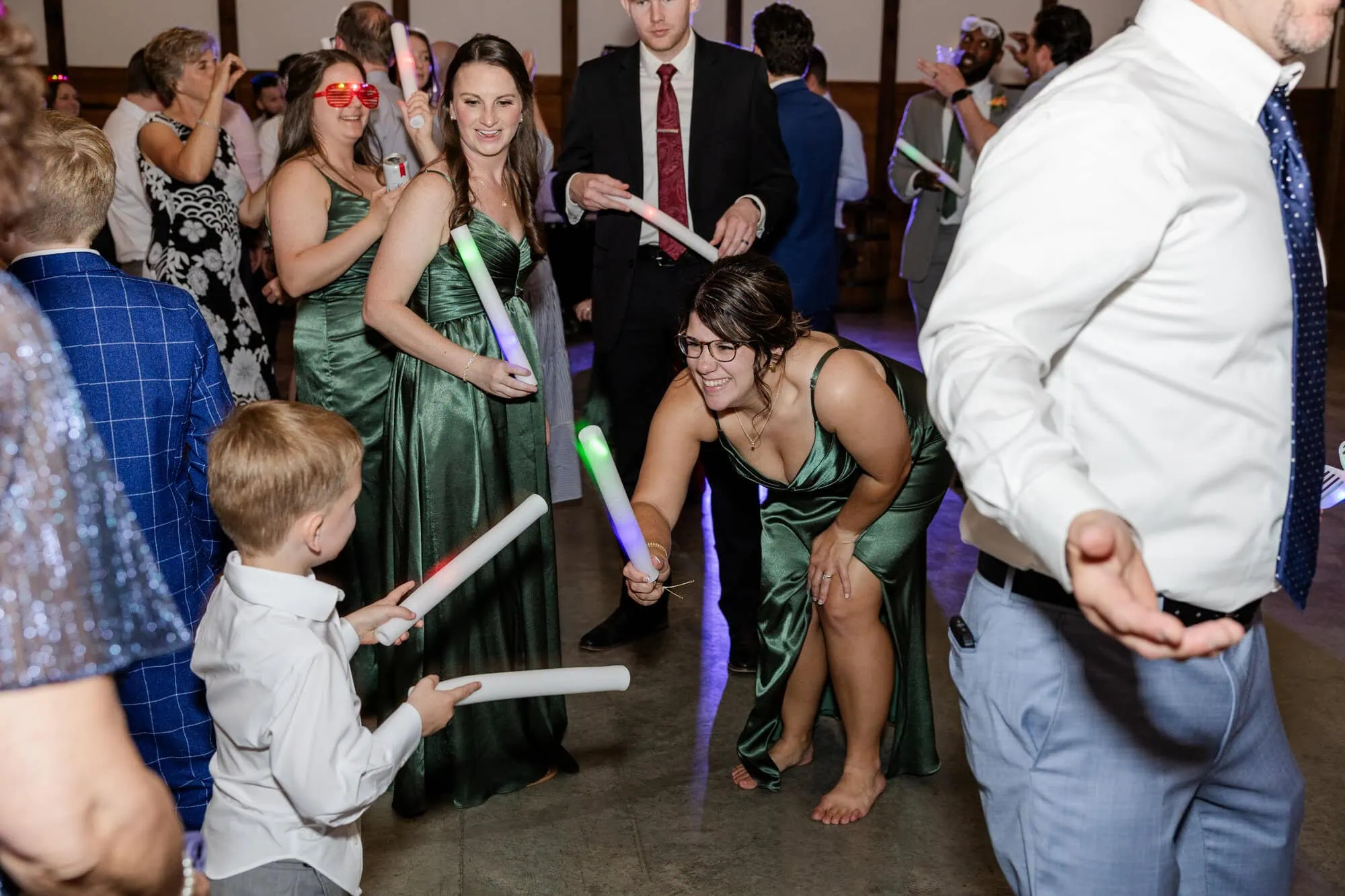 Guests in formal attire dance and play with glowing foam sticks at a lively indoor Tiffin Barn wedding. A woman in a green dress smiles at a young boy with a stick, while others socialize, capturing joyful Tiffin Barn photos throughout the night.