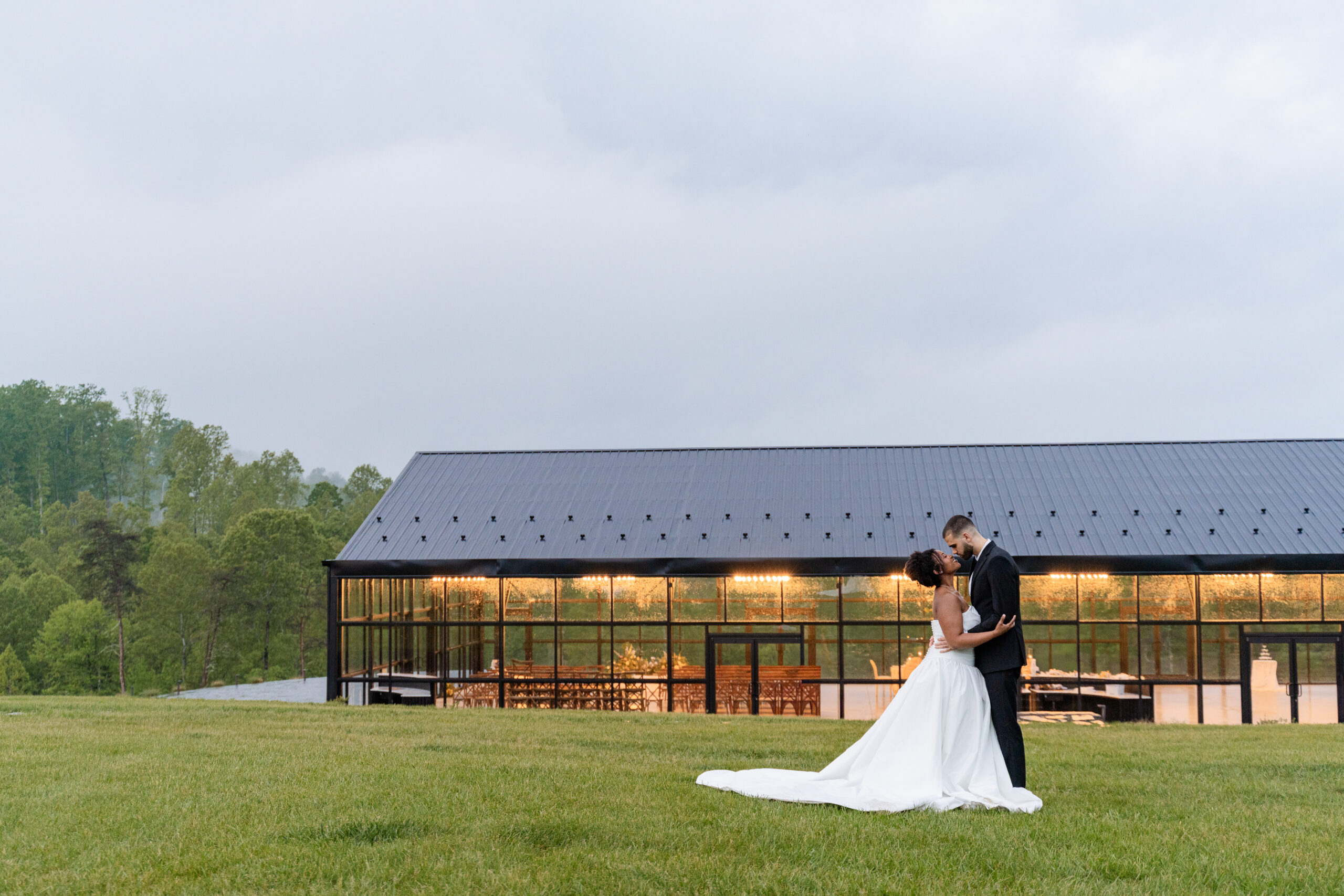 A bride and groom embrace on a grassy lawn in front of a modern barn-style venue with large windows, reminiscent of the elegant glass wedding venues popular today, with glowing lights inside under a cloudy sky.
