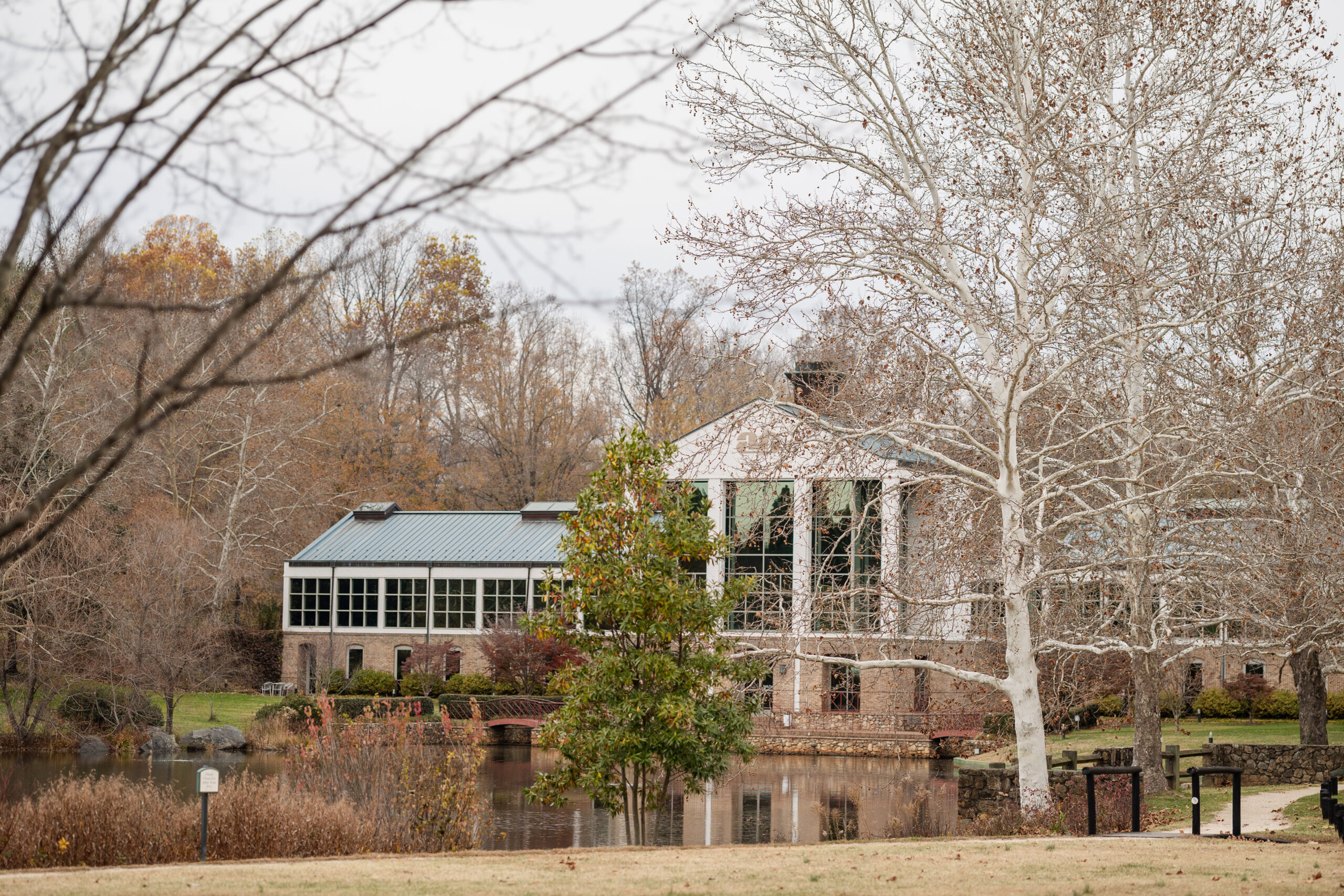 A brick building with large windows sits beside a pond, surrounded by leafless trees and autumn foliage—an elegant scene reminiscent of the serene landscapes at Boar’s Head Resort. Brown grass and a few green shrubs are in the foreground, making it easy to imagine why this could be a top choice in any thoughtful Charlottesville wedding venue review.