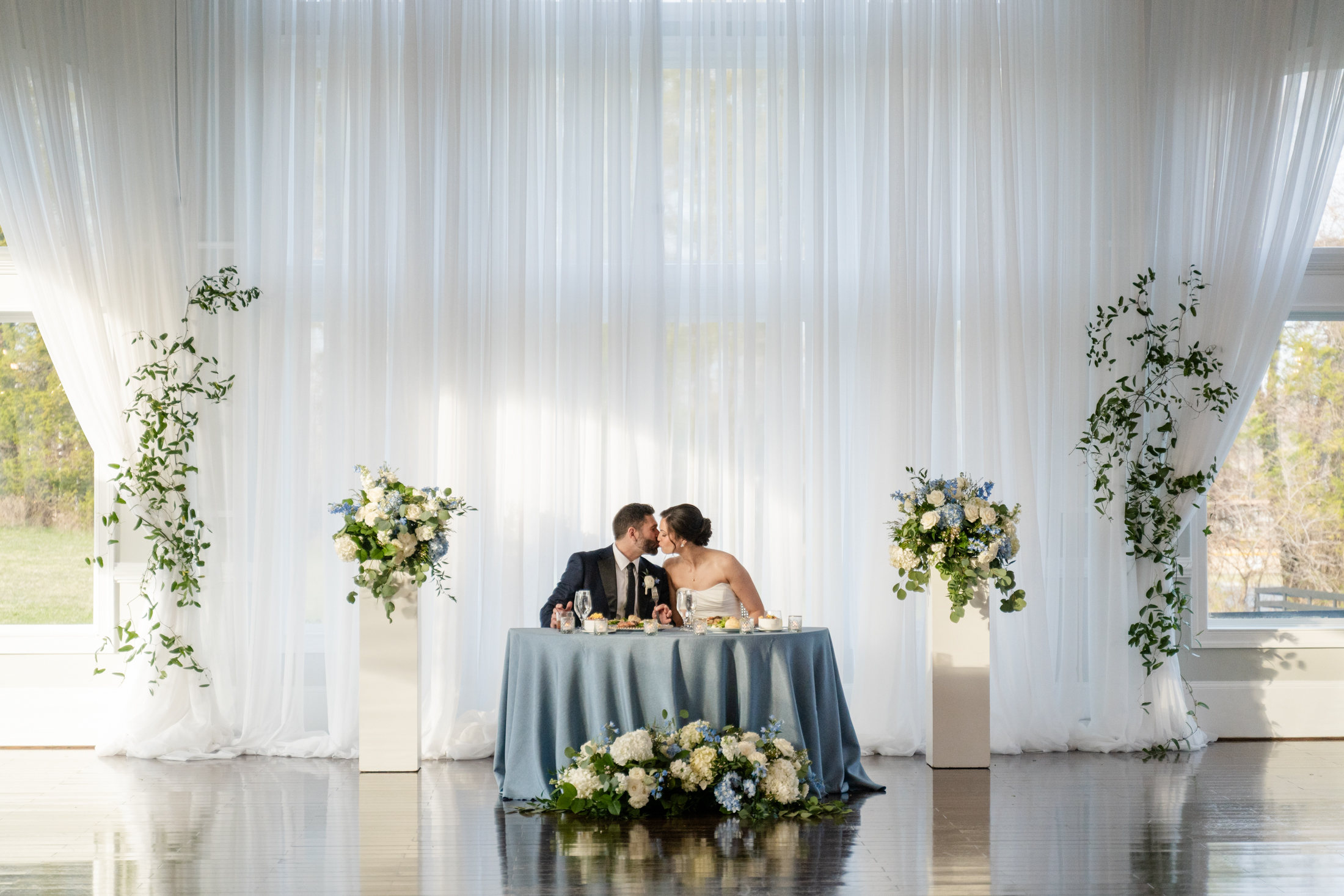 A bride and groom kiss while seated at a table with blue and white flowers, in front of large windows with white curtains and green floral arrangements on pedestals, capturing the romance of a Spring Wedding at Blackhorse Manor in Warrenton, Virginia.
