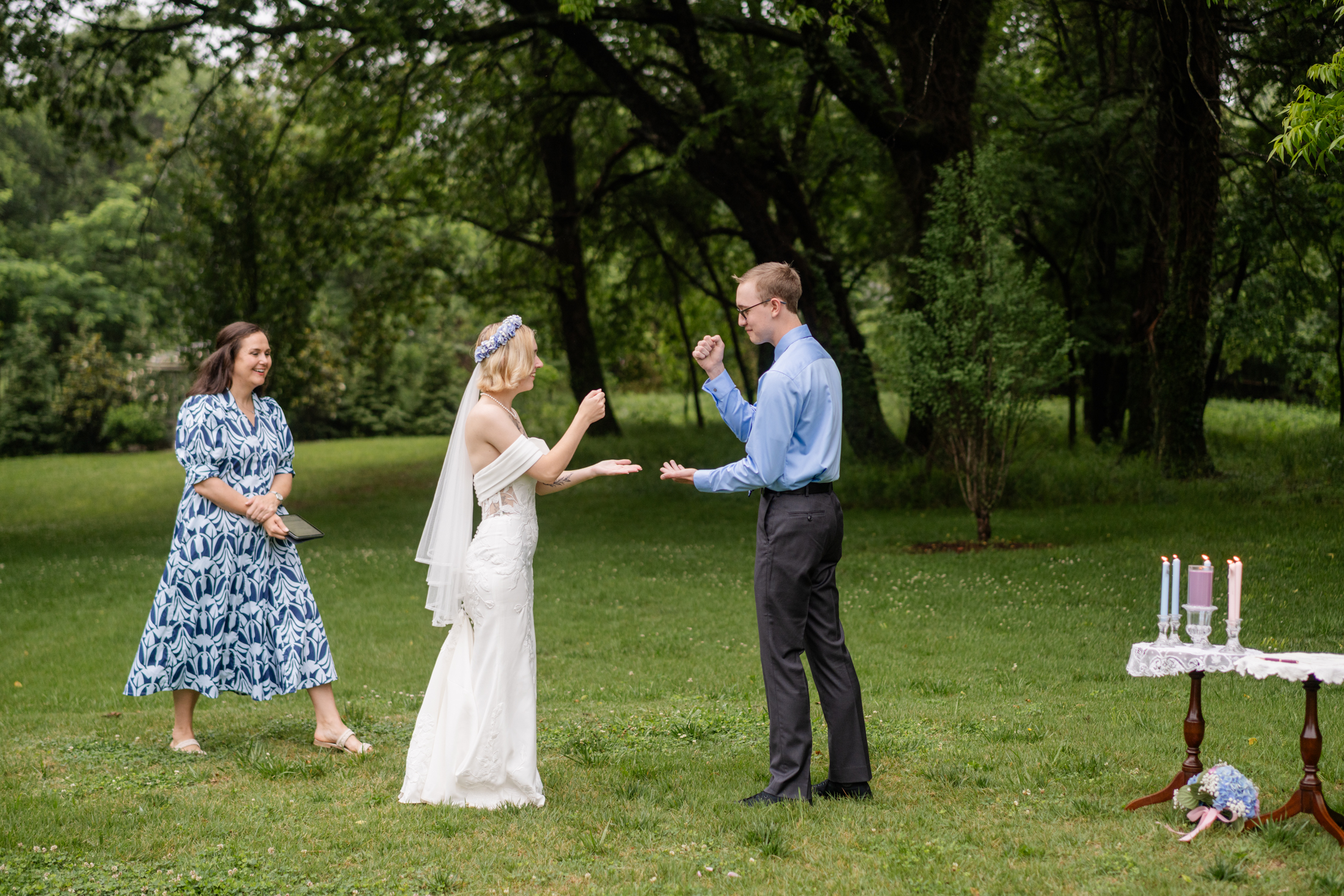 At their micro wedding at Boxwood Villa, a bride and groom play rock-paper-scissors in a grassy park with trees, while a smiling woman in a blue dress stands nearby. A small table with candles and flowers sits to the right.