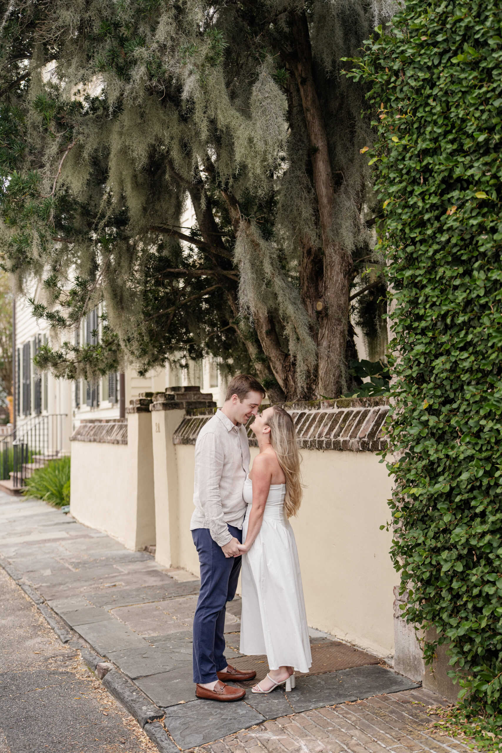 A couple stands close together on a sidewalk, smiling and touching foreheads during their Charleston engagement session, with lush greenery, a large tree, and a cream-colored building wall in the background.
