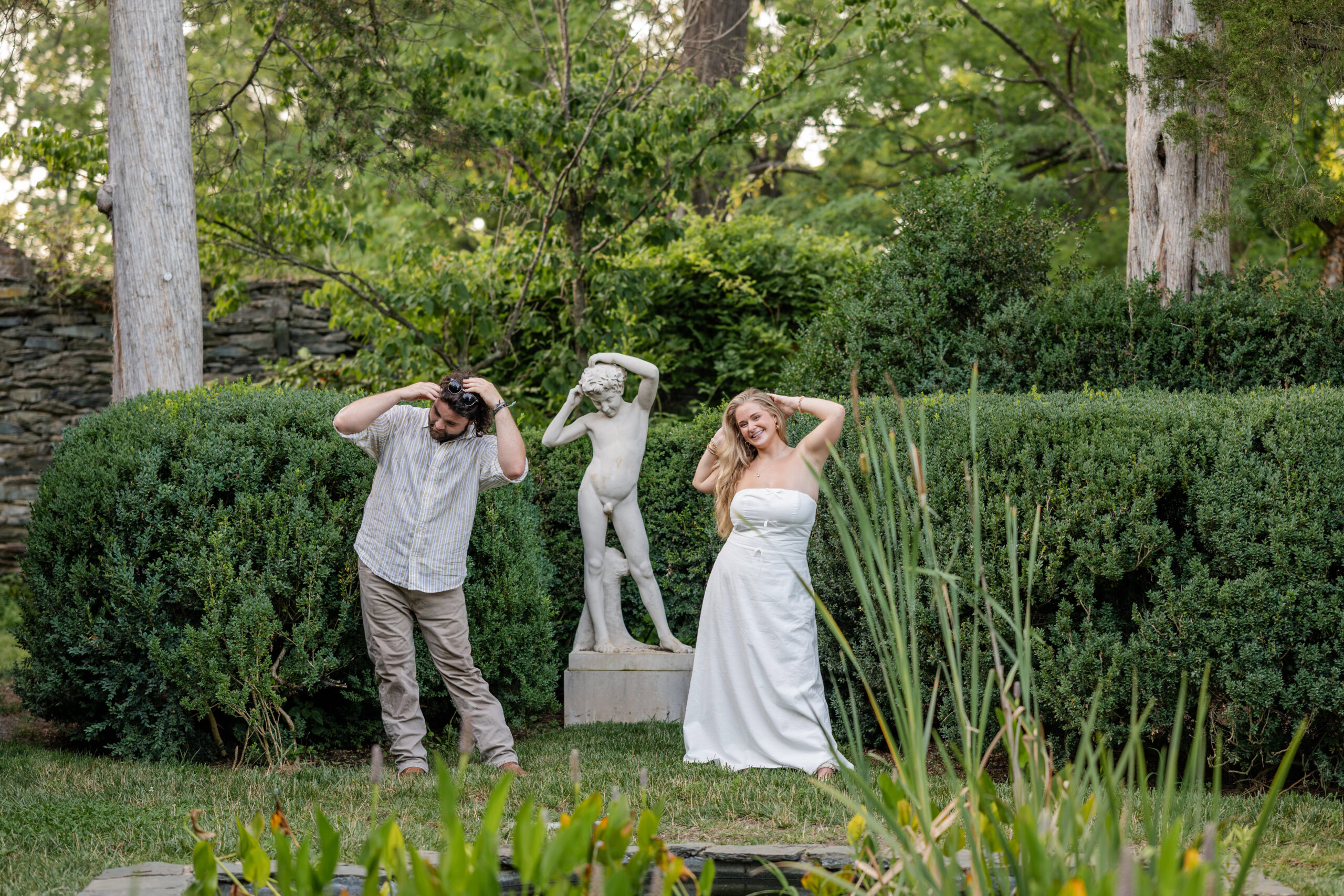 A man and woman mimic the pose of a garden statue behind them, standing in front of tall hedges and greenery during their engagement session at Oatlands. The woman wears a white dress; the man wears casual clothes.