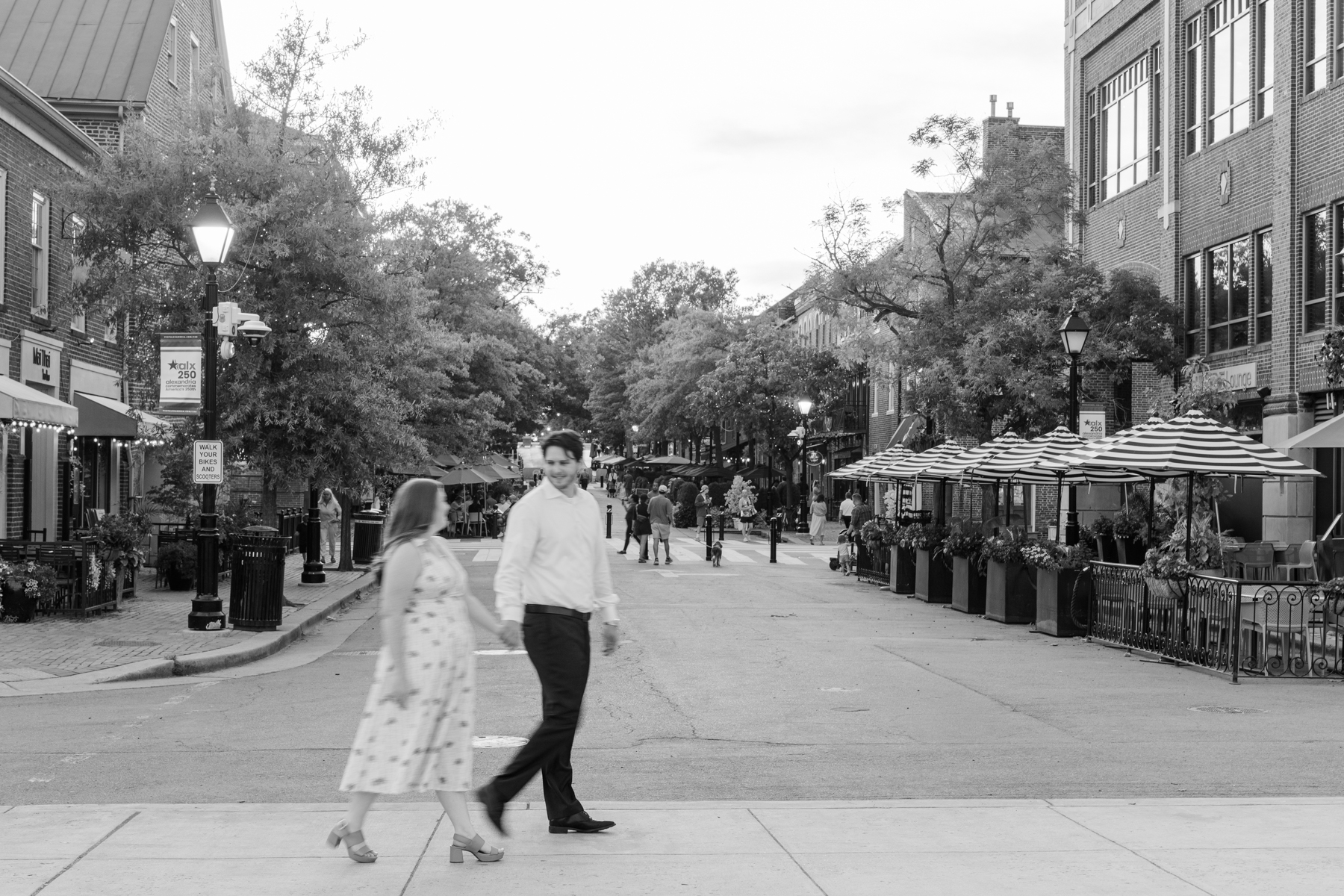 A man and woman cross a lively, tree-lined street with outdoor dining and striped umbrellas. The image is black and white, creating a classic, timeless atmosphere—perfect for Old Town Alexandria engagement photos.